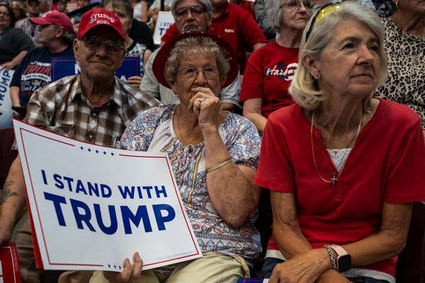 Attendees watching Mr. Trump speak at a rally, with one holding a sign that reads, “I stand with Trump.” 