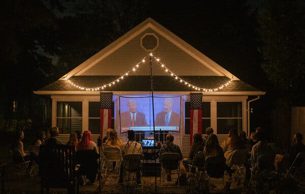 People watching a debate between Donald Trump and Joe Biden in 2020, sitting on folding chairs outside a house with a projector set up in front of it.